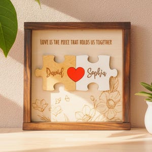 Puede incluir: Obra de arte enmarcada en madera con dos piezas de rompecabezas, una con "David" y otra con "Sophia", unidas por un corazón rojo. El texto dice "LOVE IS THE PIECE THAT HOLDS US TOGETHER". El marco es marrón oscuro.