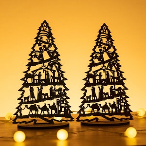 May include: Two black wooden Christmas tree ornaments with a silhouette of the nativity scene. The trees are standing on a wooden surface with white lights in the background.
