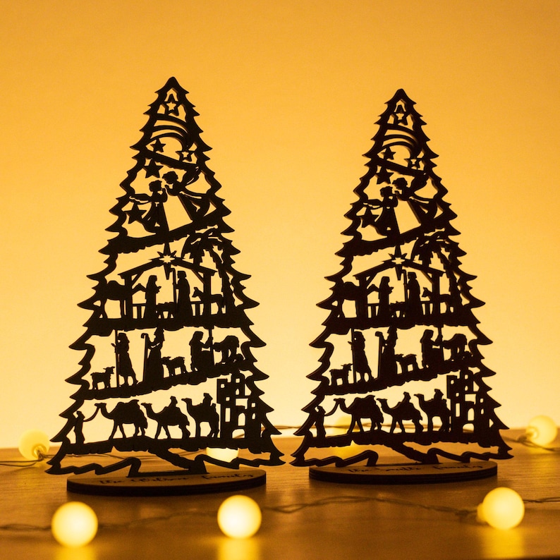 May include: Two black wooden Christmas tree ornaments with a silhouette of the nativity scene. The trees are standing on a wooden surface with white lights in the background.
