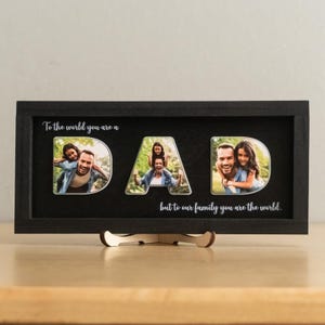 Może przedstawiać: Czarna oprawiona ekspozycja zdjęć ze słowem "DAD" zapisanym ze zdjęciami rodzinnymi. Wyświetlacz zawiera tekst "To the world you are a" nad słowem "DAD" i "but to our family you are the world" poniżej.
