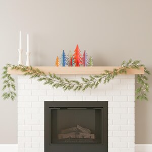 May include: A fireplace mantel decorated with a green pine garland featuring small white lights, two white taper candles in holders, and a collection of seven small, stylised wooden Christmas trees in orange, blue, red, purple, and green.
