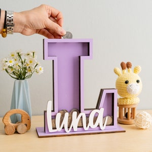 May include: Purple wooden letter L piggy bank with the name "Luna" written in white letters. A hand is placing a coin in the slot. A small stuffed giraffe and a wooden toy car are in the background.