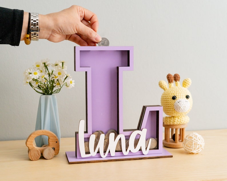 May include: Purple wooden letter L piggy bank with the name "Luna" written in white letters. A hand is placing a coin in the slot. A small stuffed giraffe and a wooden toy car are in the background.