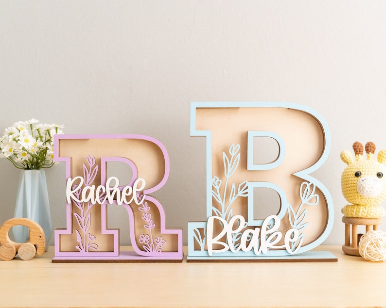 May include: Two wooden letters, one pink "R" with the name "Rachel" and one light blue "B" with the name "Blake", both with floral cutouts.