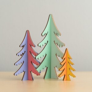 May include: A set of three freestanding tree-shaped ornaments made of flat, coloured panels. The left tree is purple and red, the centre tree is light green, and the right tree is yellow and orange. Each tree features a stylised, jagged branch design.
