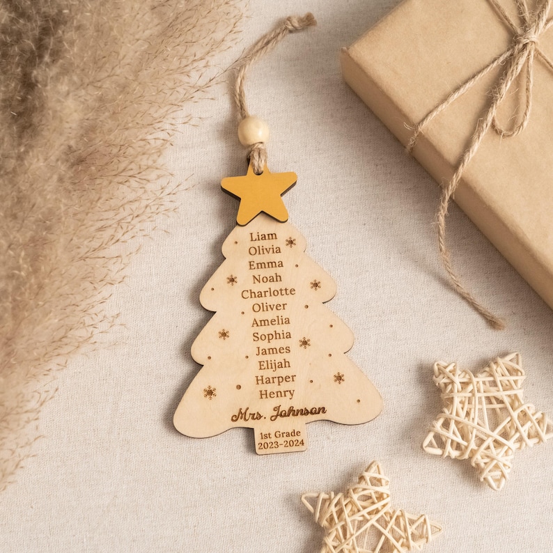 May include: Wooden Christmas tree ornament with a yellow star on top. The ornament is decorated with the names of students, "Liam, Olivia, Emma, Noah, Charlotte, Oliver, Amelia, Sophia, James, Elijah, Harper, Henry" and the text "Mrs. Johnson, 1st Grade, 2023-2024".