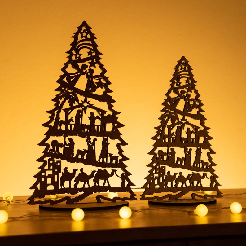 May include: Two wooden Christmas tree ornaments with a silhouette of the nativity scene. The trees are brown and the silhouettes are black. The trees are on a table with white lights.