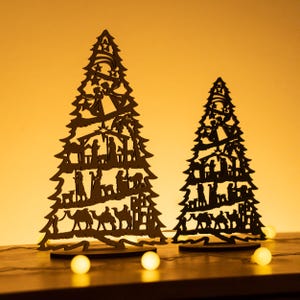 May include: Two wooden Christmas tree ornaments with a silhouette of the nativity scene. The trees are black and stand on a wooden base. The background is a warm yellow.