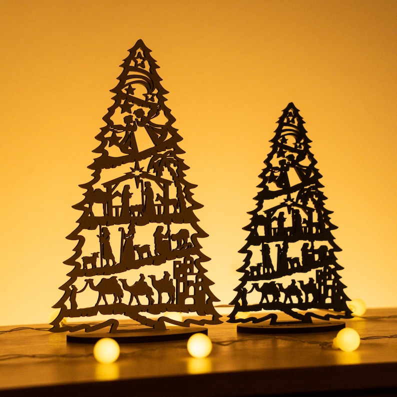 May include: Two wooden Christmas tree ornaments with a silhouette of the nativity scene. The trees are black and stand on a wooden base. The background is a warm yellow.