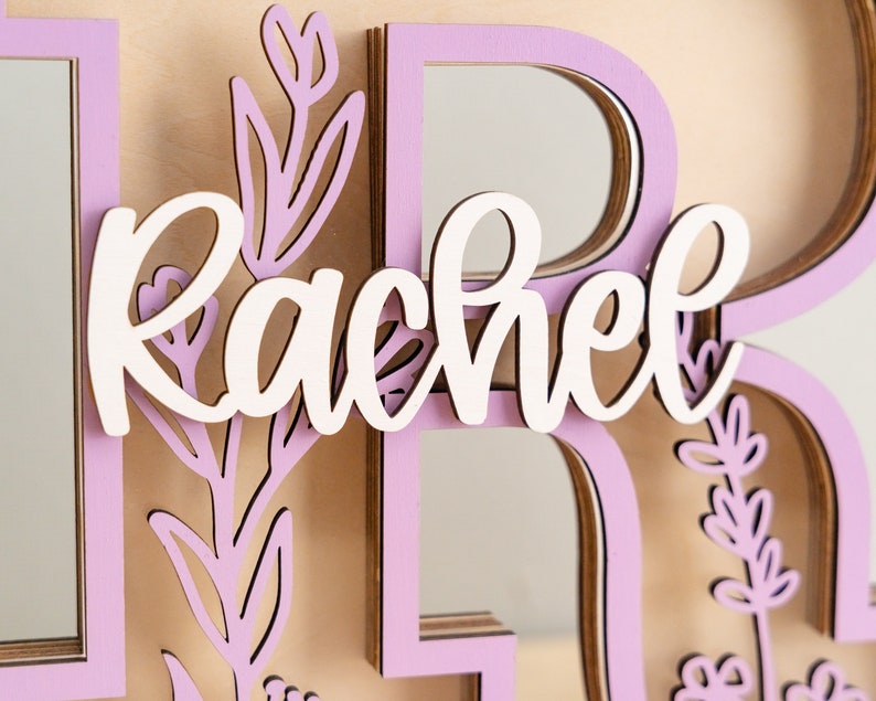 May include: A wooden sign with the name "Rachel" in white script lettering. The sign is decorated with pink floral cutouts and has a layered, 3D effect.