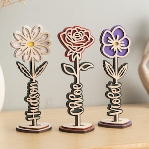 May include: Three wooden flower decorations with personalized names. The flowers are a white daisy, a red rose, and a purple violet. Each flower has a wooden stem with the name "Hudson", "Grace", and "Violet" written on it.