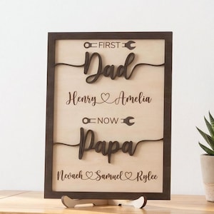 Puede incluir: Cartel enmarcado en madera con las palabras "First Dad" y "Now Papa" en escritura marrón oscuro. También se muestran los nombres Henry, Amelia, Nevach, Samuel y Rylee. El letrero está apoyado en un soporte de madera.