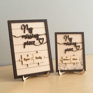 May include: Two wooden signs with the text "My missing piece" and a heart. The signs have a brown frame and a light wood background. The larger sign has the names "Ryan" and "Emily" on two puzzle pieces. The smaller sign has the names "Adrian" and "Olivia" on two puzzle pieces.