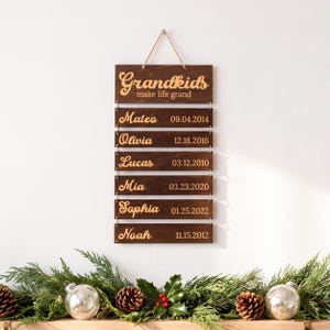 May include: A dark brown wooden wall hanging sign with six rectangular slats connected by metal rings. The top slat reads "Grandkids make life grand" and the five lower slats feature names and dates: Mateo 09.04.2014, Olivia 12.18.2016, Lucas 03.12.2010, Mia 03.23.2020, Sophia 01.25.2022, and Noah 11.15.2012.
