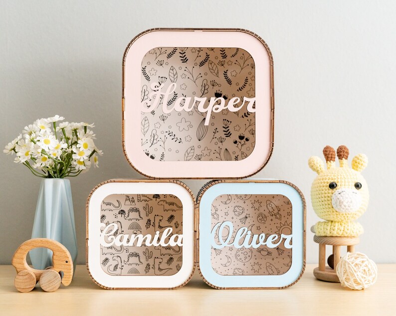 Puede incluir: Tres cajas de sombra cuadradas de madera con marcos de colores claros en rosa, blanco y azul. Cada caja cuenta con un panel frontal transparente con un nombre personalizado en letra blanca: Harper, Camila y Oliver. Los fondos muestran patrones de flores, dinosaurios y temas espaciales.