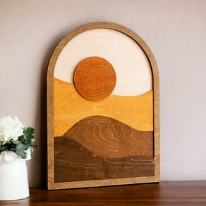 Pu&ograve; includere: Opera d'arte da parete in legno con un design paesaggistico a strati con un sole e colline. L'opera utilizza tonalit&agrave; di marrone, beige e arancione, incorniciata in un arco. Il pezzo &egrave; esposto su una superficie di legno scuro.