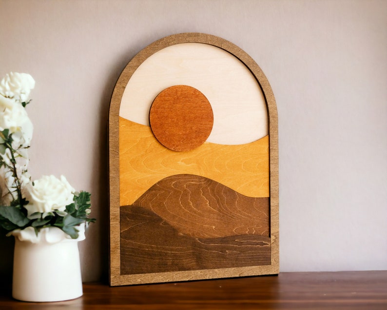 Pu&ograve; includere: Opera d'arte da parete in legno con un design paesaggistico a strati con un sole e colline. L'opera utilizza tonalit&agrave; di marrone, beige e arancione, incorniciata in un arco. Il pezzo &egrave; esposto su una superficie di legno scuro.
