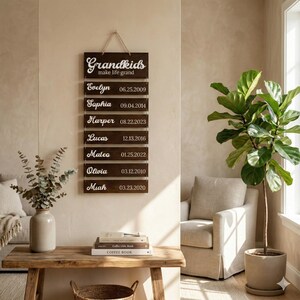 May include: A vertical wall hanging sign made of dark brown wood planks connected by twine. The top plank reads "Grandkids make life grand" in white script. Below are seven planks featuring names and birth dates: Evelyn, Saphia, Harper, Lucas, Mateo, Olivia, and Miah.