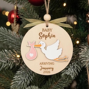 May include: A wooden Christmas ornament with the name "Sophia" and the text "Baby" above it. A stork carrying a pink bundle is depicted. The text "Arriving January 2026" is below. The ornament is hanging on a Christmas tree.