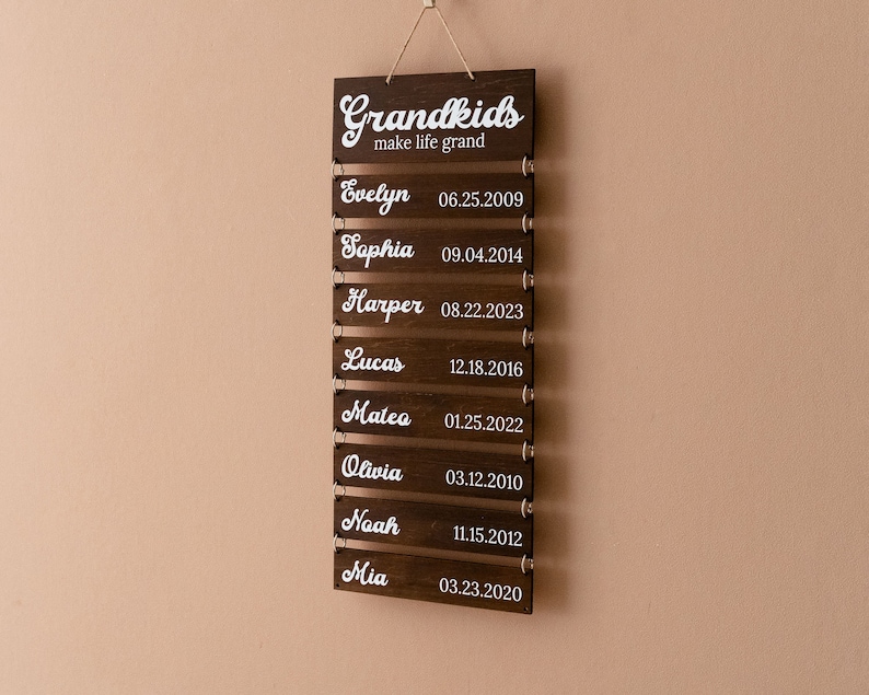 May include: A dark brown wooden wall hanging sign featuring the white text Grandkids make life grand at the top, followed by eight individual horizontal slats, each displaying a name and a birth date in white script and numerical font.