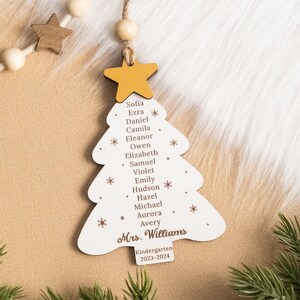 May include: White wooden Christmas tree ornament with a yellow star on top. The ornament has a list of names, including Sofia, Ezra, Daniel, Camila, Eleanor, Owen, Elizabeth, Samuel, Violet, Emily, Hudson, Hazel, Michael, Aurora, and Avery. The ornament also includes the text "Mrs. Williams", "Kindergarten", and "2023-2024".