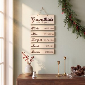 May include: A vertical wooden wall hanging featuring the text "Grandkids make life grand" on the top plaque, followed by five smaller plaques listing the names Olivia, Mia, Harper, Noah, and Lucas with their respective birth dates in dark brown.