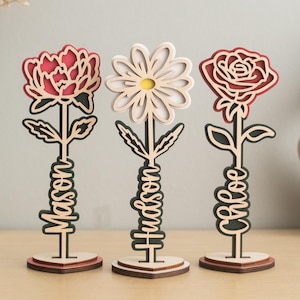 May include: Three wooden flower sculptures with personalized names. The flowers are a red rose, a white daisy, and a red peony. Each flower is on a wooden base.