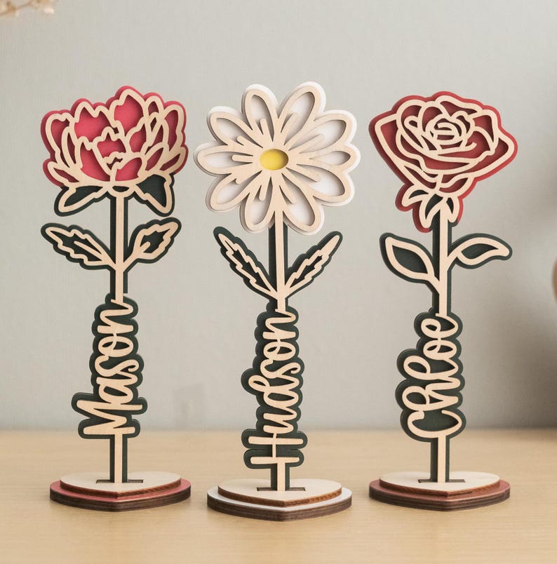 May include: Three wooden flower sculptures with personalized names. The flowers are a red rose, a white daisy, and a red peony. Each flower is on a wooden base.