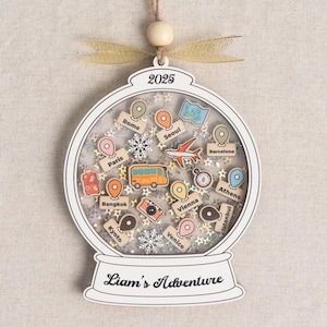 May include: A white wooden snow globe ornament with the year 2025 at the top. Inside are travel-themed wooden cutouts of a bus, airplane, and landmarks. The base reads "Liam's Adventure".