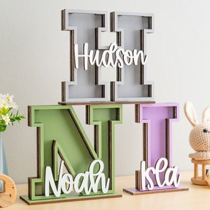 May include: Three wooden letters, H, N, and I, painted in gray, green, and purple, with the names "Hudson", "Noah", and "Isla" written in white on the front of each letter.