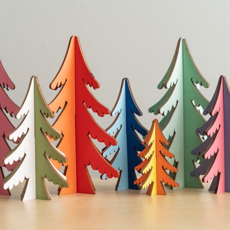May include: A collection of freestanding, flat-cut wooden tree ornaments in various sizes and colours, including red, orange, light green, dark green, blue, yellow, and purple, featuring jagged, stylised branch silhouettes and visible dark edges.