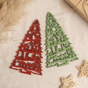 May include: Two wooden Christmas tree ornaments, one red and one green, with a nativity scene design. The ornaments are on a beige fabric background.