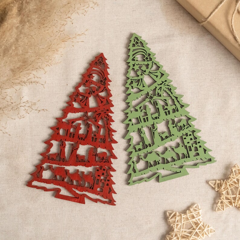 May include: Two wooden Christmas tree ornaments, one red and one green, with a nativity scene design. The ornaments are on a beige fabric background.