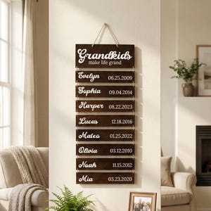May include: A dark brown wooden wall hanging sign featuring the text Grandkids make life grand at the top, followed by eight horizontal slats listing names and birth dates including Evelyn 06.25.2009, Sophia 09.04.2014, Harper 08.22.2023, Lucas 12.18.2016, Mateo 01.25.2022, Olivia 03.12.2010, Noah 11.15.2012, and Mia 03.23.2020.