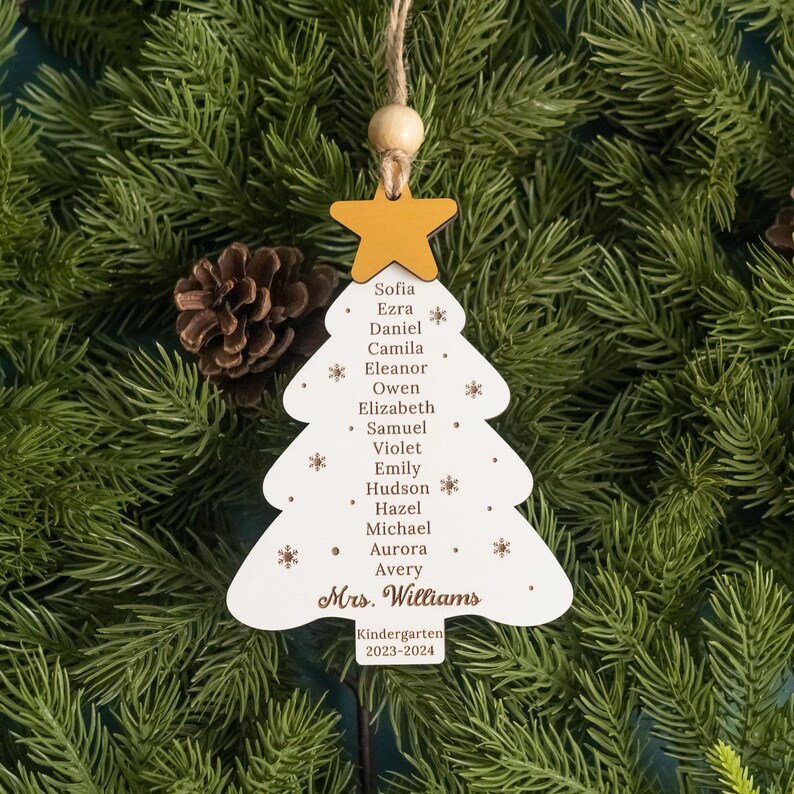 May include: White wooden Christmas tree ornament with a yellow star on top. The ornament has the names of children and the text "Mrs. Williams Kindergarten 2023-2024" written on it.