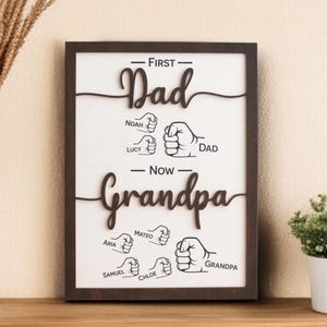May include: A framed wooden sign with the words "First Dad" and "Now Grandpa" in a dark brown script. The names Noah, Lucy, Aria, Mateo, Samuel, and Chloe are listed below the words. The sign also features illustrations of fists.