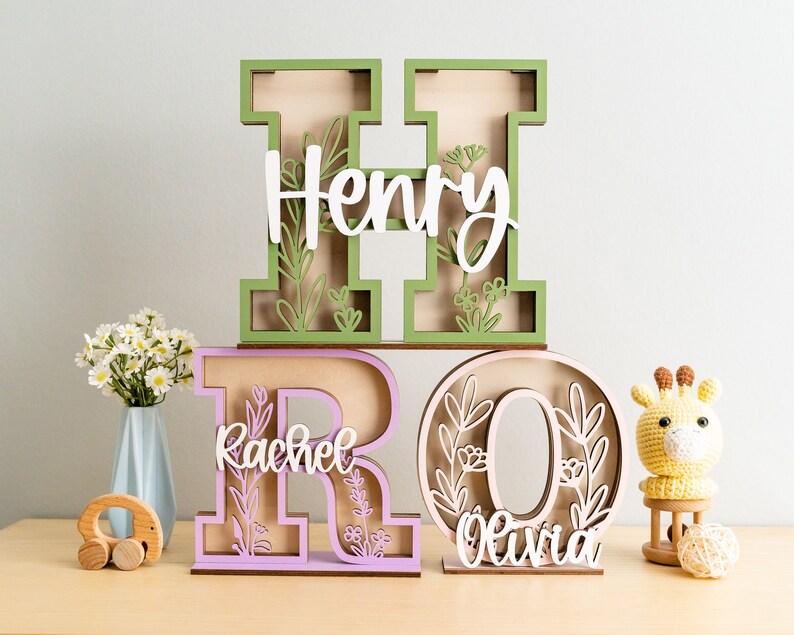 May include: Wooden letters with floral designs spelling out the names Henry, Rachel, and Olivia. The letters are painted in green, purple, and natural wood tones.