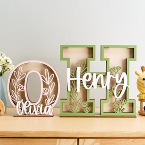 May include: Wooden letters spelling "Olivia" and "Henry" in pink and green with floral cutouts. The letters are standing on a wooden surface.