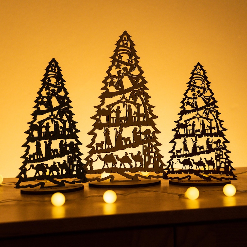 May include: Three black wooden Christmas tree ornaments with a nativity scene design. The trees are standing on a table with warm yellow lights.