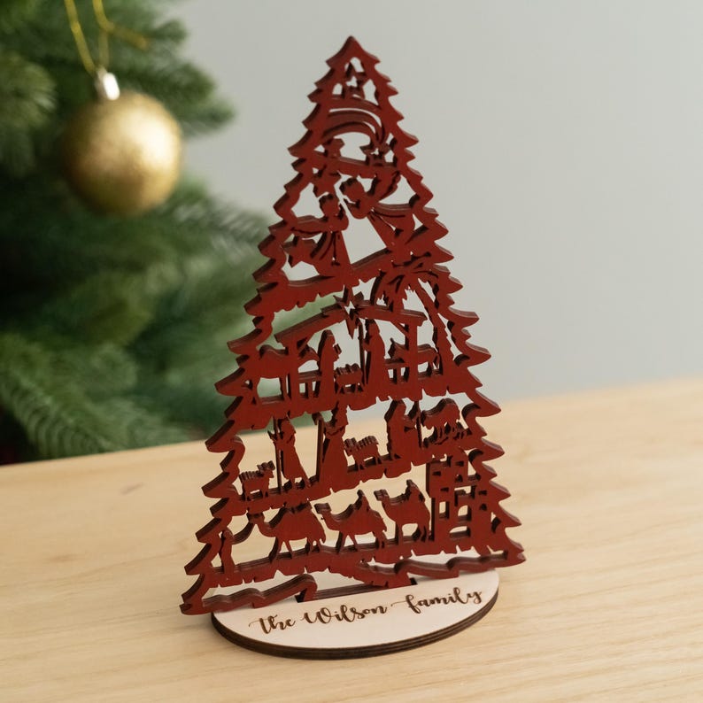 May include: Red wooden Christmas tree ornament with a nativity scene cutout. The base of the tree is personalized with the text "The Wilson Family".