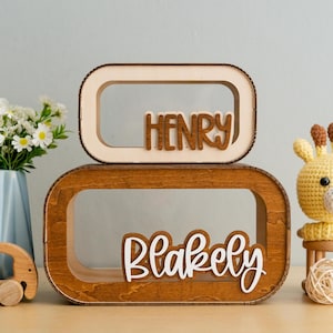 May include: Two wooden name plaques with rounded edges. The top plaque is light wood with the name "HENRY" in dark wood letters. The bottom plaque is dark wood with the name "Blakely" in white letters.