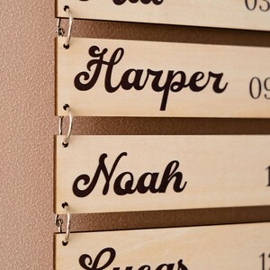 May include: A series of rectangular light-colored wooden plaques connected by silver metal rings. Each plaque features a name printed in a dark brown cursive font, specifically the names Mia, Harper, Noah, and Lucas, with partial numbers visible.