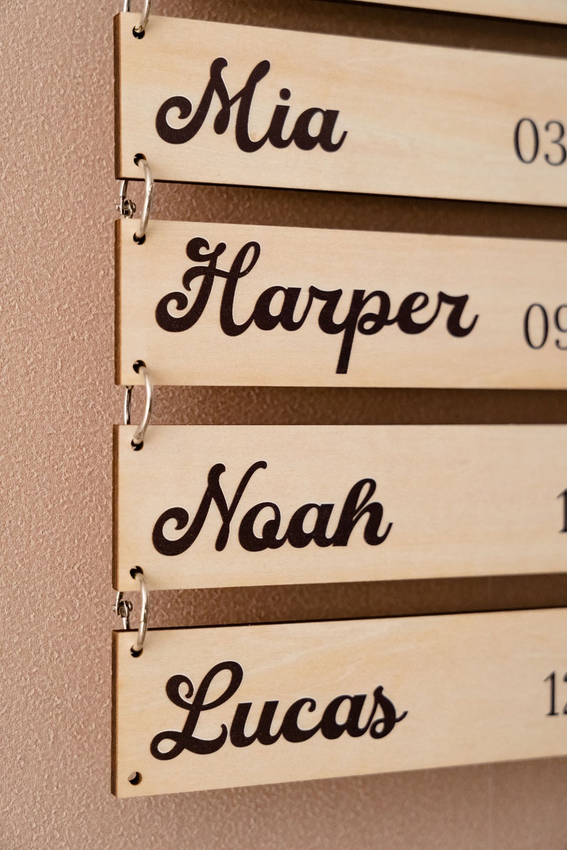 May include: A series of rectangular light-colored wooden plaques connected by silver metal rings. Each plaque features a name printed in a dark brown cursive font, specifically the names Mia, Harper, Noah, and Lucas, with partial numbers visible.
