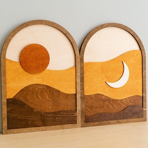 Pu&ograve; includere: Due opere d'arte da parete in legno raffiguranti un paesaggio. Uno presenta un sole, l'altro una luna crescente. L'opera d'arte utilizza sfumature di marrone, arancione e beige, con un design a strati. Le cornici sono arcuate.