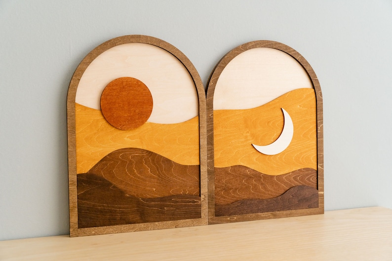 Pu&ograve; includere: Due opere d'arte da parete in legno raffiguranti un paesaggio. Uno presenta un sole, l'altro una luna crescente. L'opera d'arte utilizza sfumature di marrone, arancione e beige, con un design a strati. Le cornici sono arcuate.