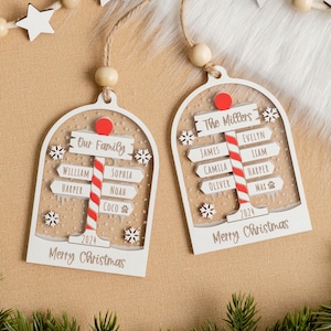 May include: Two arched white wooden Christmas ornaments featuring a central red and white striped pole with directional signs. The signs are engraved with names and the text Our Family, The Millers, and Merry Christmas, with 2024 and snowflakes.