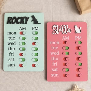 May include: Two pet schedule boards, one mint green and one pink. The mint green board has the name "Rocky" and a dog silhouette. The pink board has the name "Stella" and a cat silhouette. Each board has a weekly schedule with AM and PM toggles.