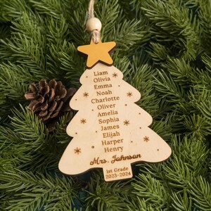 May include: A wooden Christmas tree ornament with a yellow star on top. The ornament is decorated with the names of students and the teacher's name, "Mrs. Johnson", along with the grade and year. The ornament is surrounded by green pine branches.