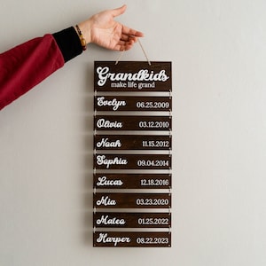 May include: A dark brown wooden wall hanging sign featuring the white text Grandkids make life grand at the top, followed by eight smaller rectangular wooden slats connected by metal rings, each displaying a name and a birth date in white script.