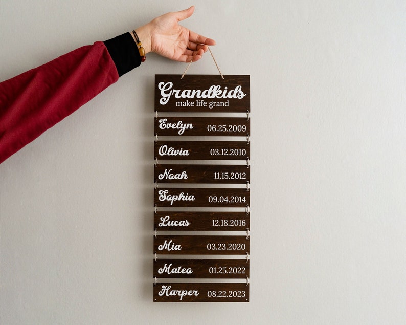 May include: A dark brown wooden wall hanging sign featuring the white text Grandkids make life grand at the top, followed by eight smaller rectangular wooden slats connected by metal rings, each displaying a name and a birth date in white script.